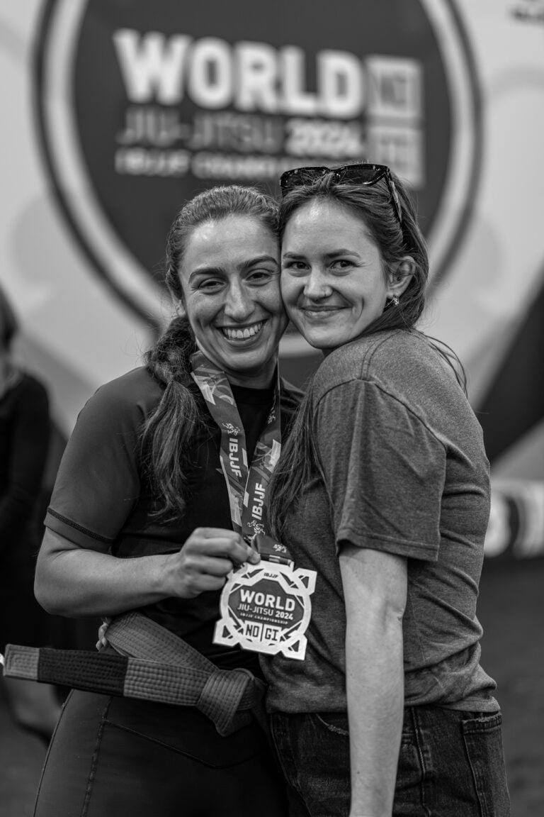Two women hugging and smiling, holding an IBJJF medal from No-Gi Worlds.