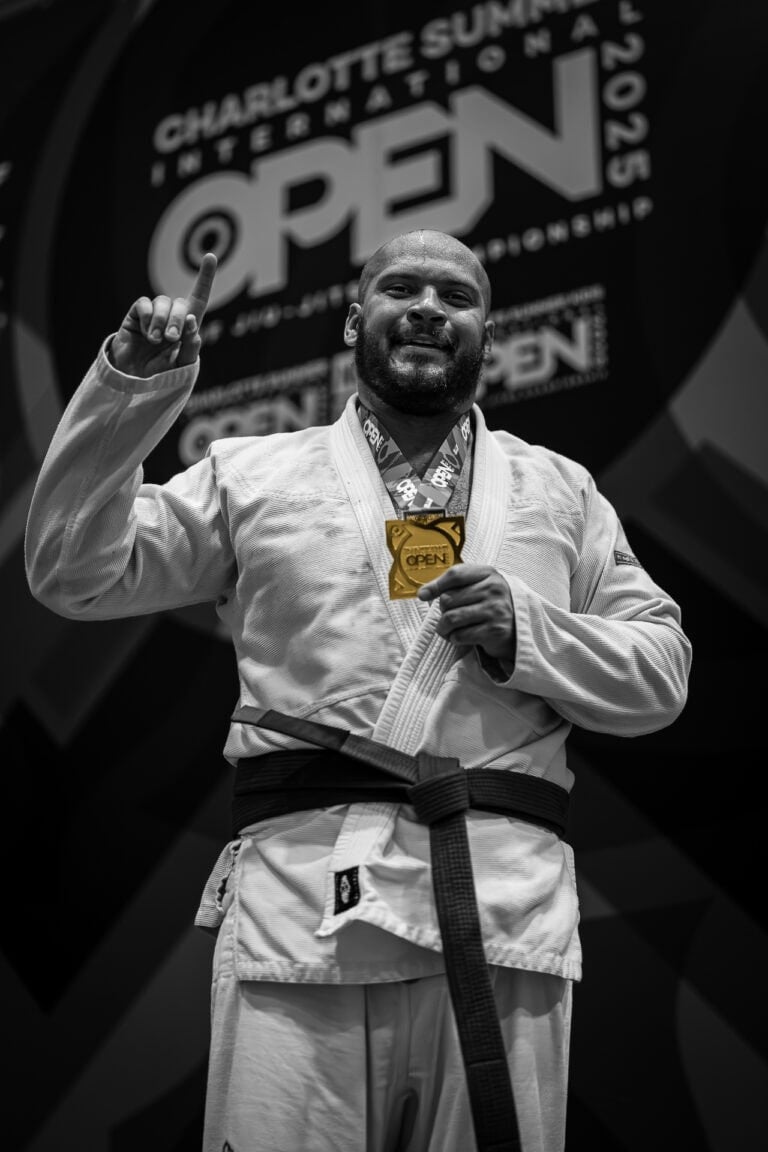 Martial artist in gi with black belt holds gold medal and raises index finger at competition.