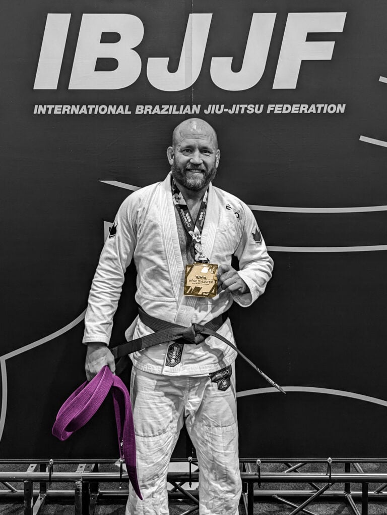 Man in Brazilian jiu-jitsu gi and black belt holding purple belt and Gold Wolds medal at IBJJF event.
