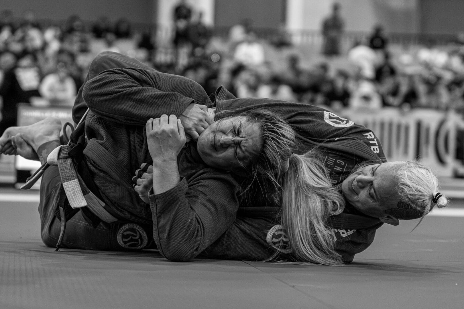 Two female martial artists competing in a Brazilian Jiu-Jitsu match with audience in background