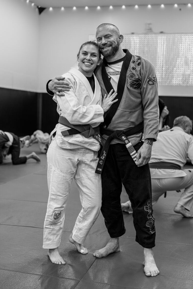 Two martial artists in gi uniforms and black belts smiling together in a training dojo.