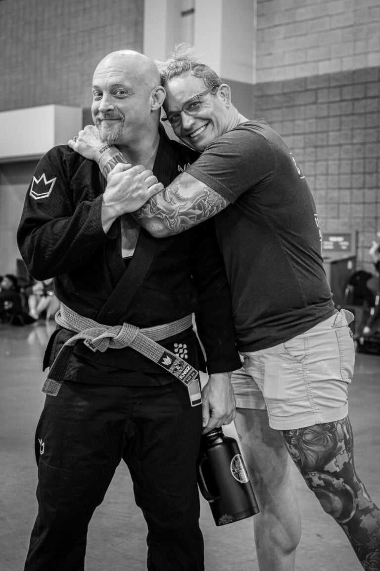 Two people indoors, one in a martial arts gi with gray belt, the other in t-shirt and shorts with tattoos.