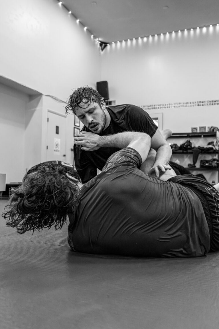 Two men grappling on a padded floor in a martial arts training facility in black and white.