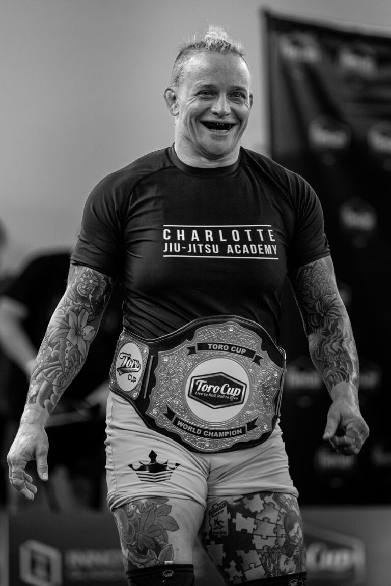 Tattooed athlete wearing Charlotte Jiu-Jitsu Academy gear with Toro Cup World Champion belt.