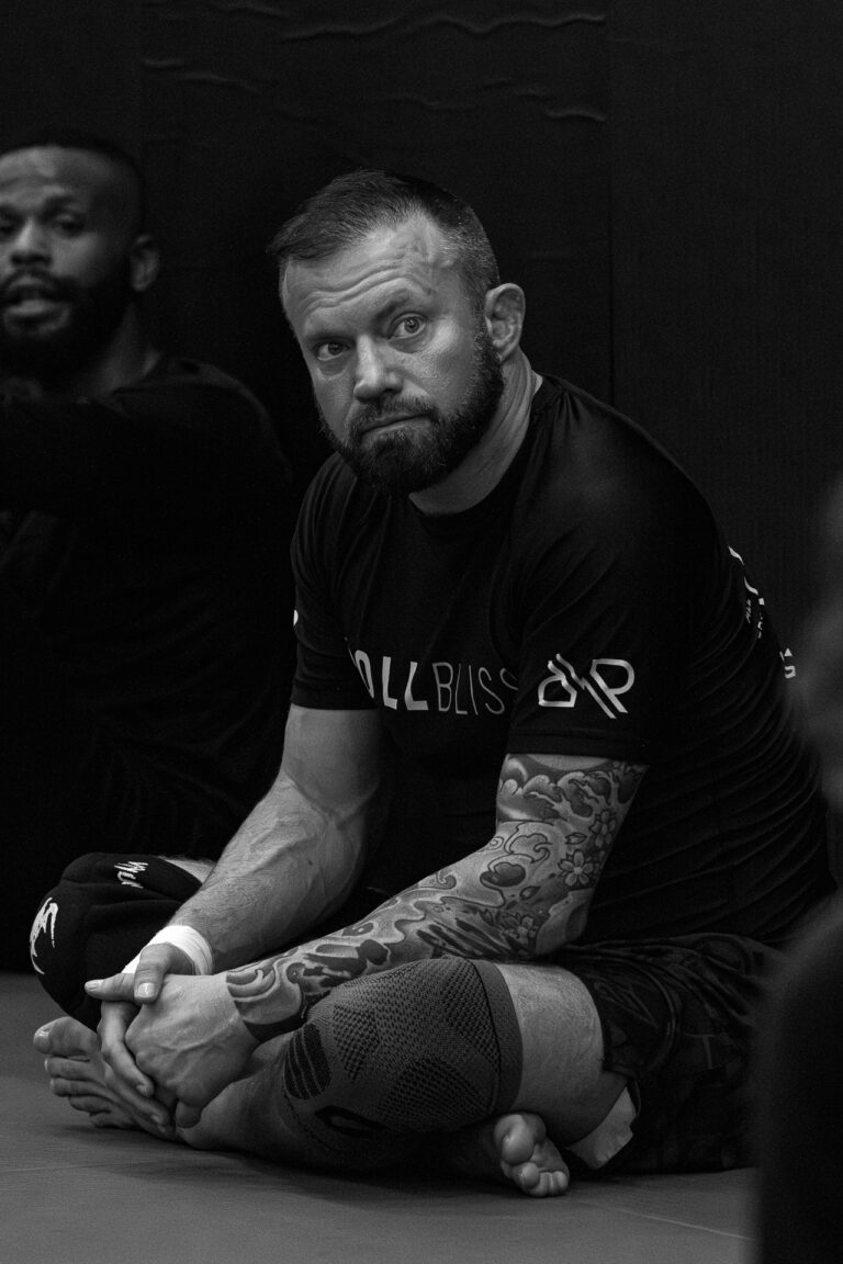 Man with tattoos and knee brace sitting cross-legged on the floor in a training setting.