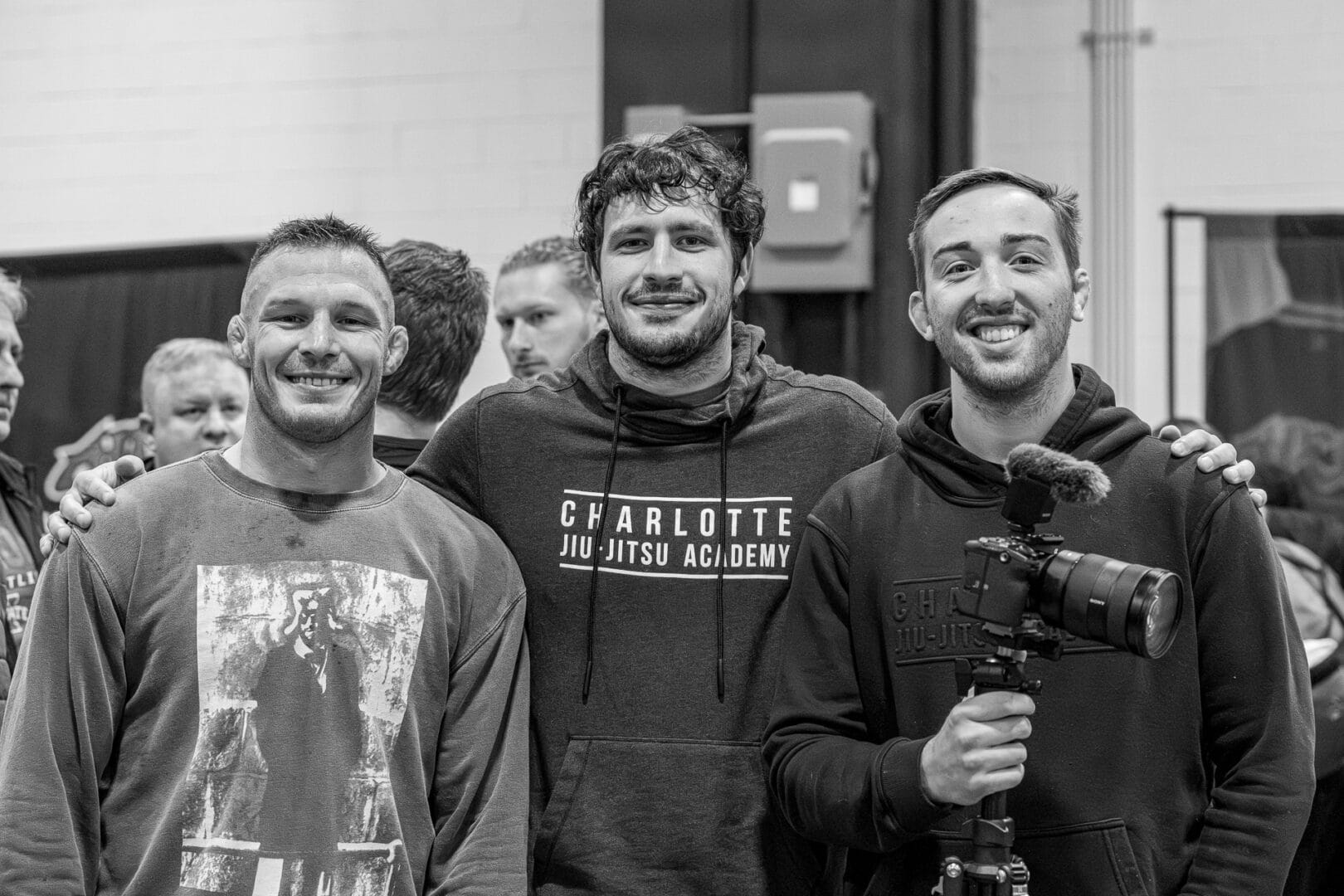 Three men indoors smiling, one holding a camera, middle wearing Charlotte Jiu Jitsu Academy hoodie.